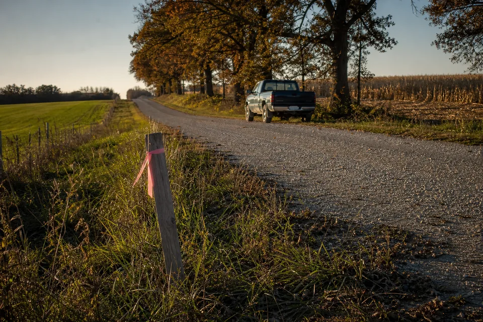 Rural land parcel with a mix of pasture, a tree line and a harvested crop field behind a gravel county road and a wooden survey stake