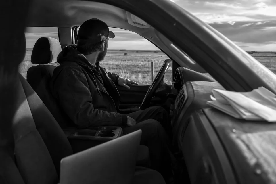 Land investor in a pickup truck reviewing a stack of mail offer letters and a laptop beside rural acreage