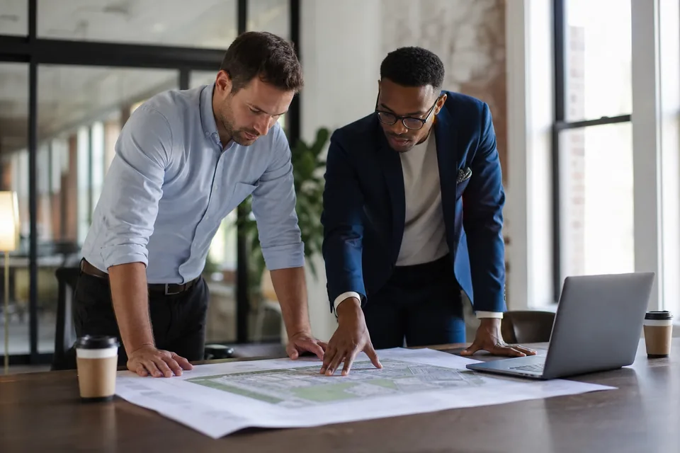 Two developers at a conference table reviewing a printed site plan and parcel map next to a laptop