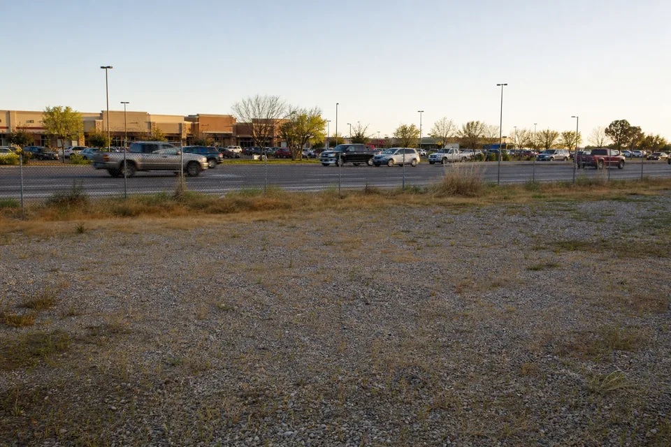 Vacant commercial infill lot with a chain link fence beside a busy suburban arterial road and strip retail